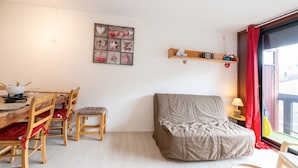 A cozy room with a wooden dining table, chairs, a cushioned bench, a wall-mounted shelf with decorative items, and a large window with red curtains. - Le Corbier - Studio avec belle vue montagne (Villarembert)