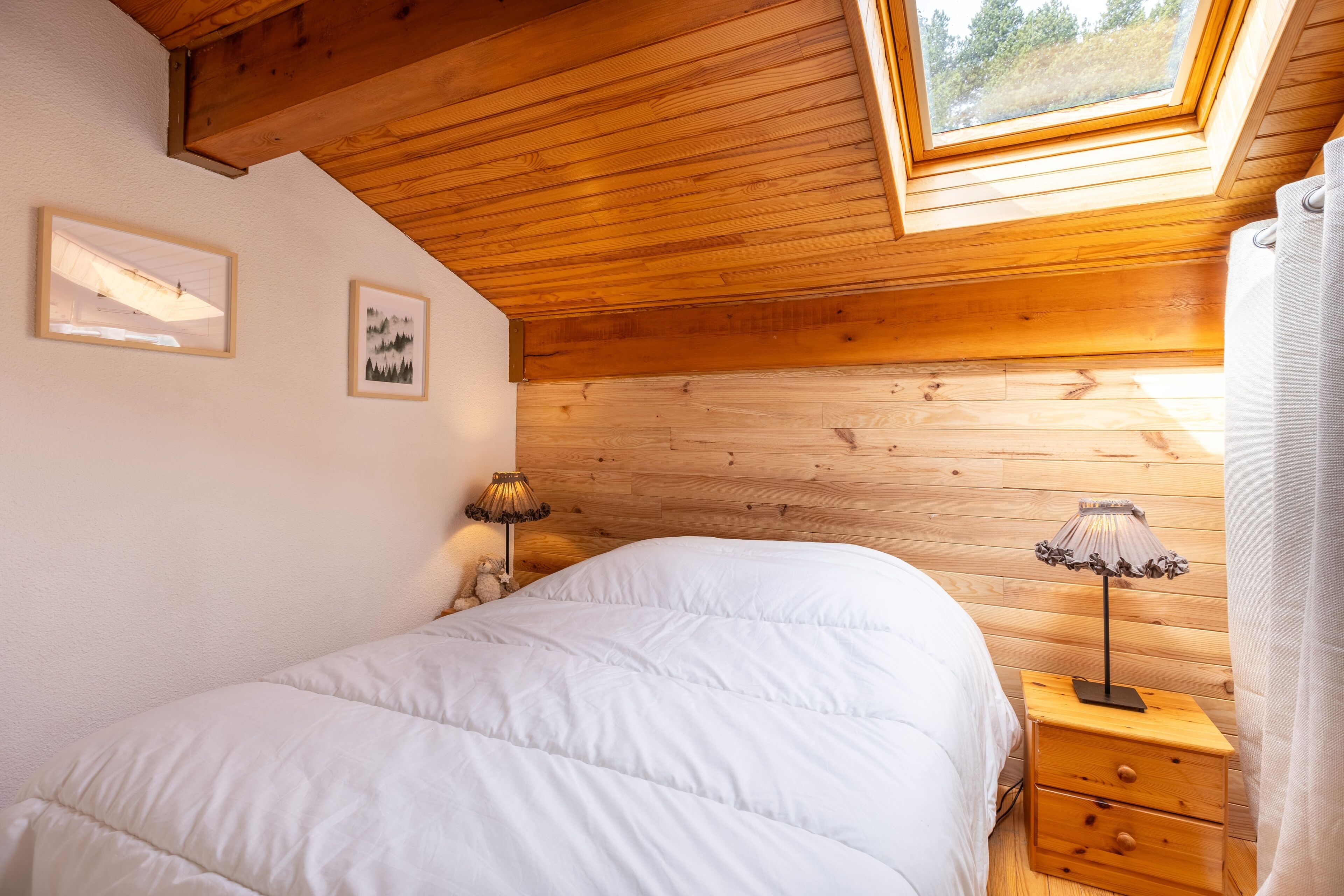 Une chambre avec un plafond en bois, une lucarne, un lit avec une literie blanche, deux tables de chevet avec des lampes et des tableaux encadrés accrochés au mur.