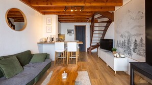 Una sala acogedora con un sofá verde, una mesa de centro de madera y un televisor. Se aprecia un comedor con sillas y una mesa, así como una escalera que lleva a otra habitación.