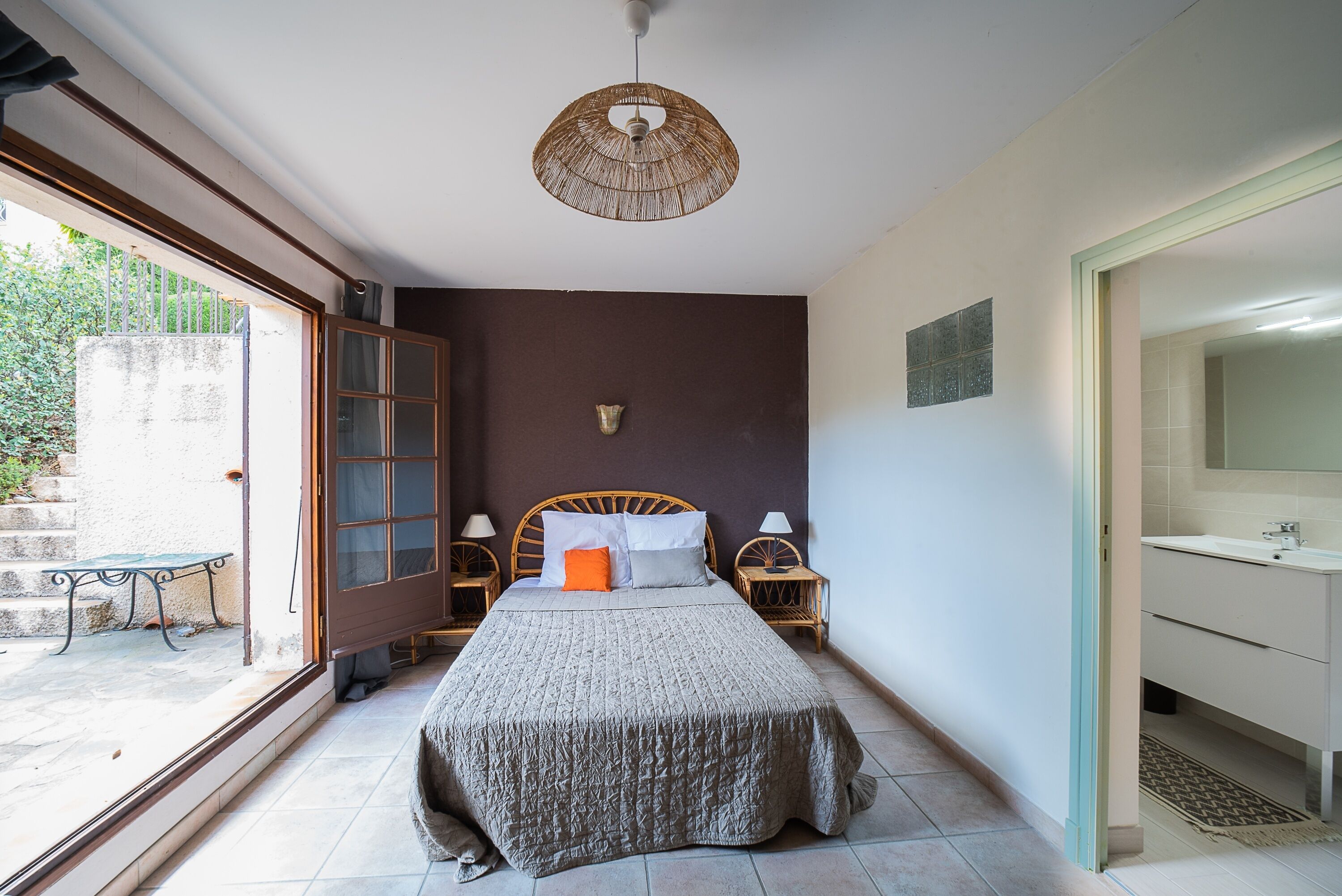 Une chambre à coucher comprenant un lit, des tables de chevet, un luminaire suspendu, un banc à l’extérieur, ainsi qu’une salle de bain avec un lavabo et un miroir.