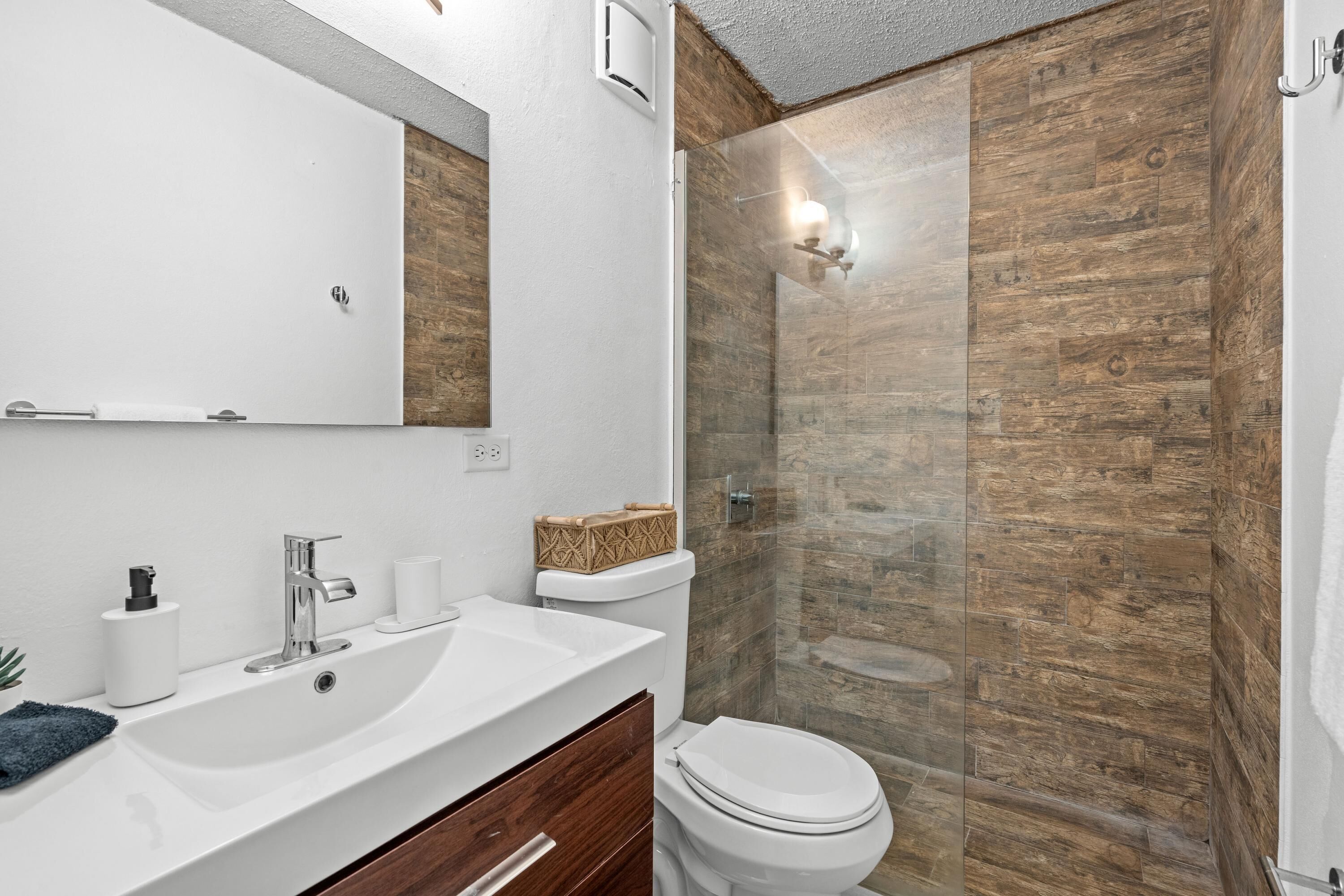 A modern bathroom with a white sink, mirror, and toilet. It features a wooden wall and a glass shower enclosure.