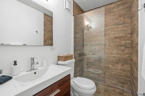 A modern bathroom with a white sink, mirror, and toilet. It features a wooden wall and a glass shower enclosure. - Otium Ideal Marina Getaway Balcony Parking (Fajardo)