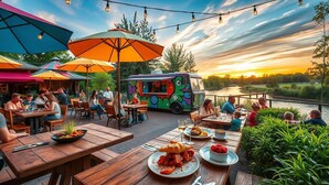 An outdoor dining area with wooden tables and colorful umbrellas, a food truck, and a scenic view of a lake at sunset.