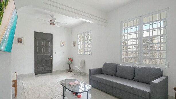 A spacious living room with a grey sofa, a glass coffee table, a ceiling fan, and large windows with white frames.