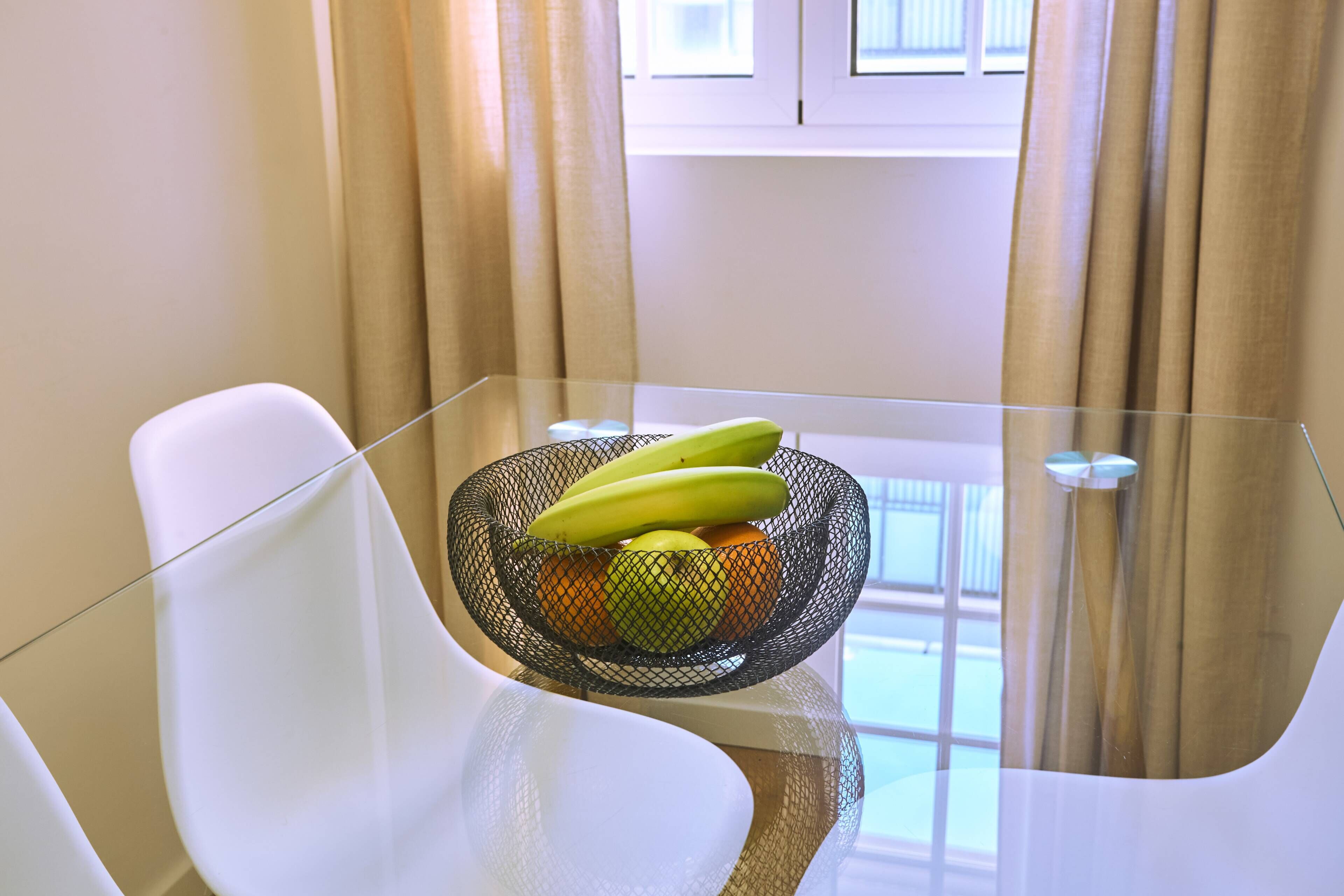A glass table with a fruit bowl containing bananas and apples, white chairs, and beige curtains.