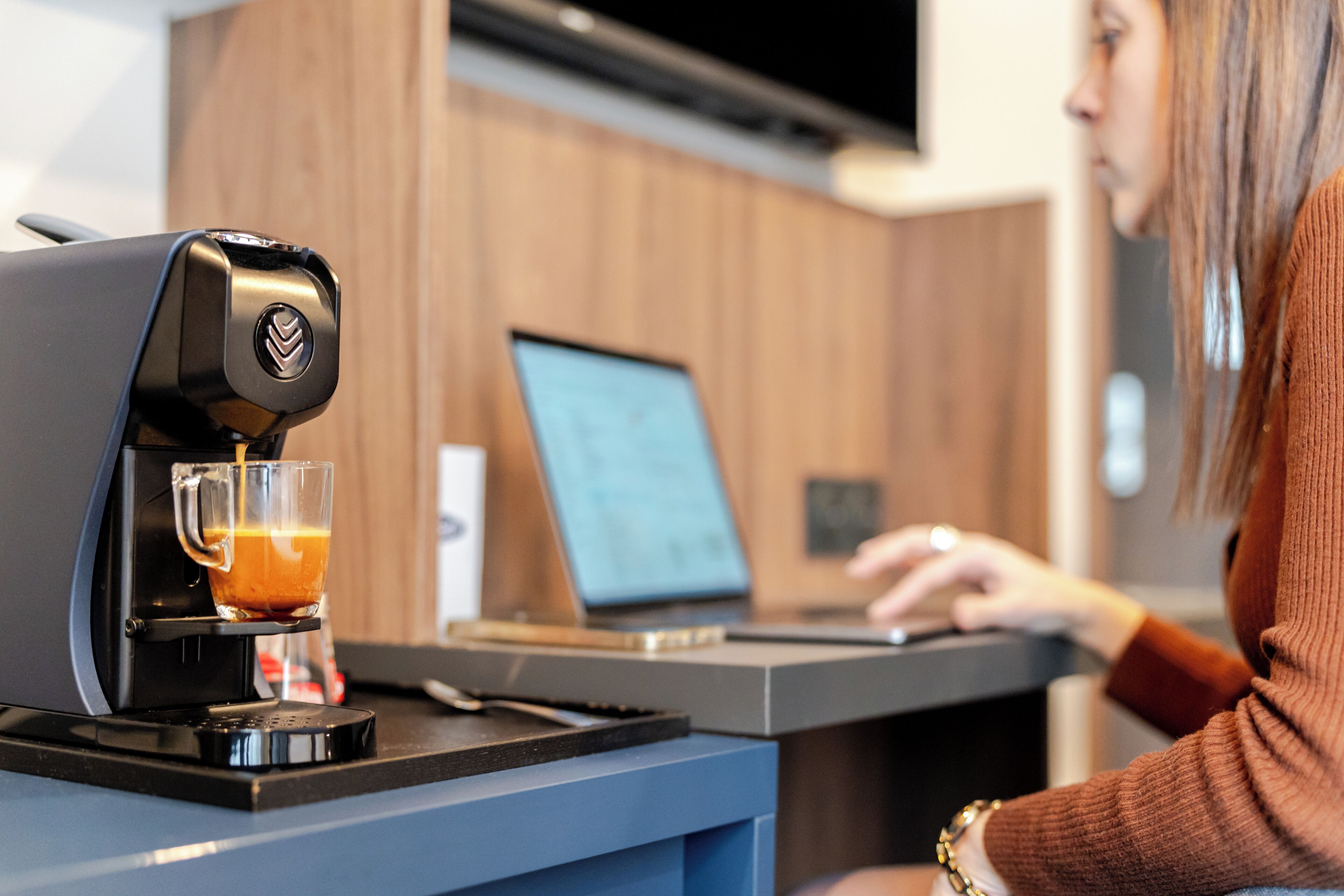 Máquina de café a servir café num copo, um laptop sobre uma secretária e uma pessoa a trabalhar no laptop.