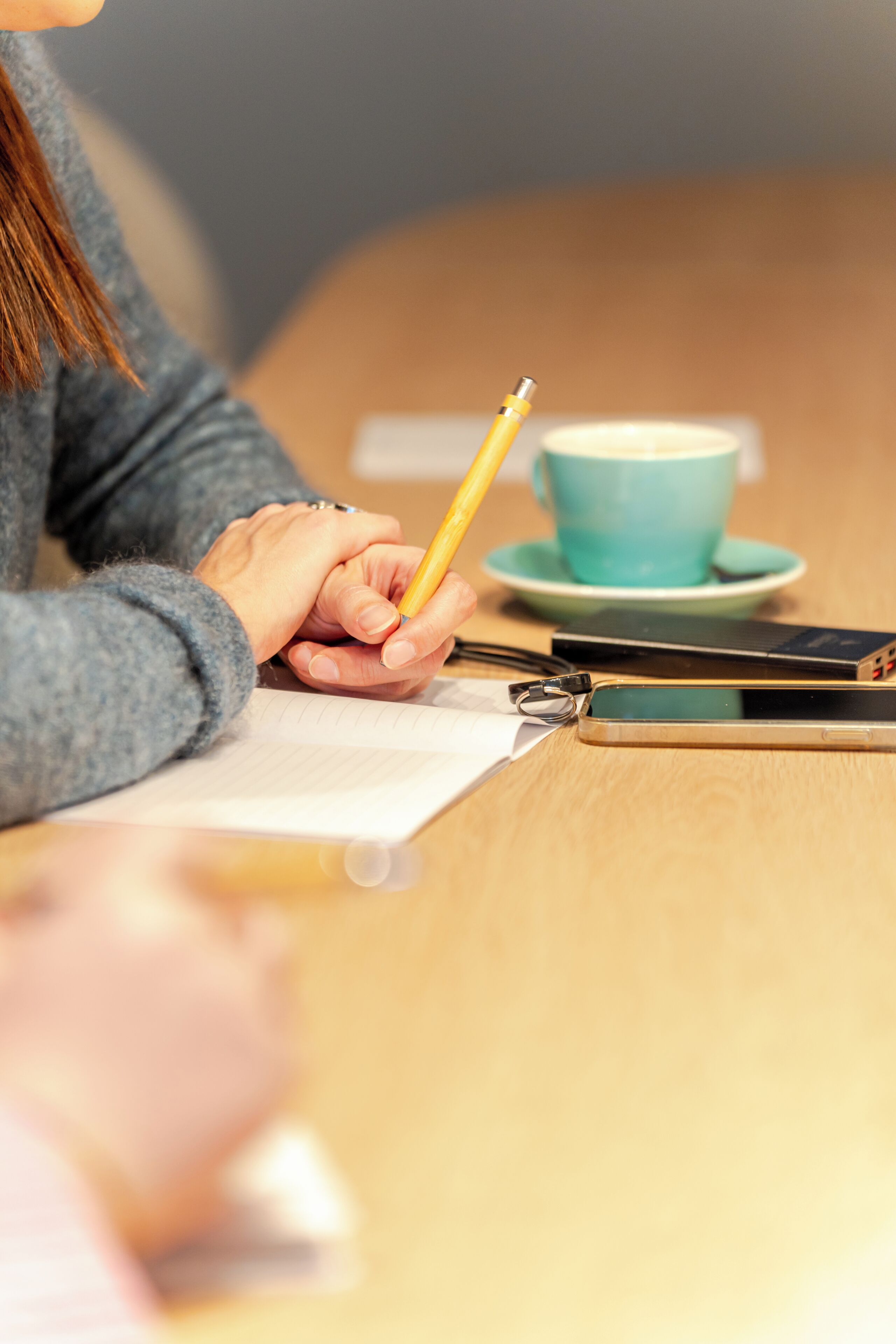 Una persona escribe con un lápiz en un bloc, un teléfono inteligente y una taza sobre una mesa.