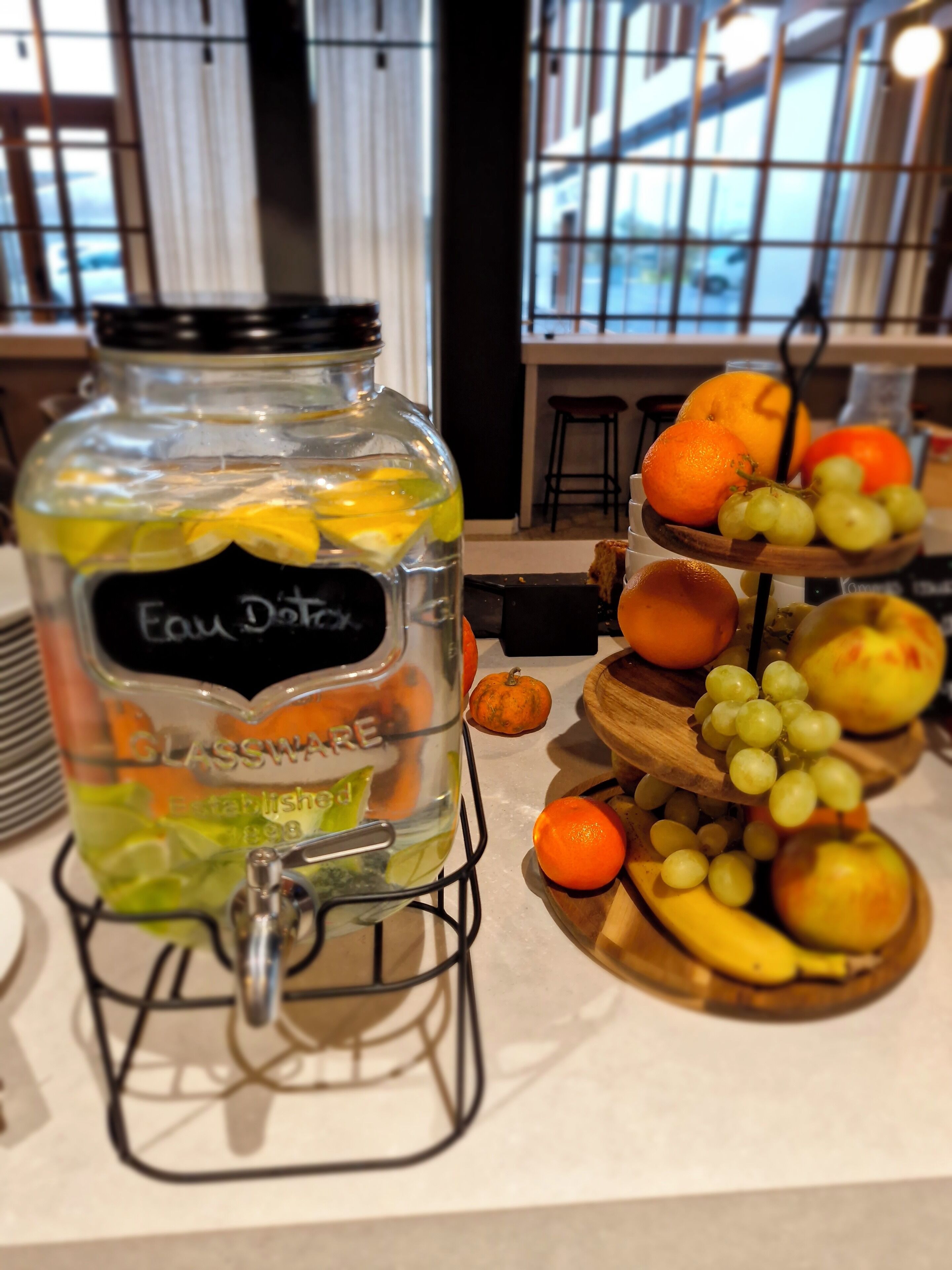 Un bocal en verre étiqueté « Fau Docks » avec une étiquette noire, rempli de tranches d’agrumes et muni d’un robinet en métal. À côté, un présentoir en bois expose des oranges, des raisins et une citrouille.