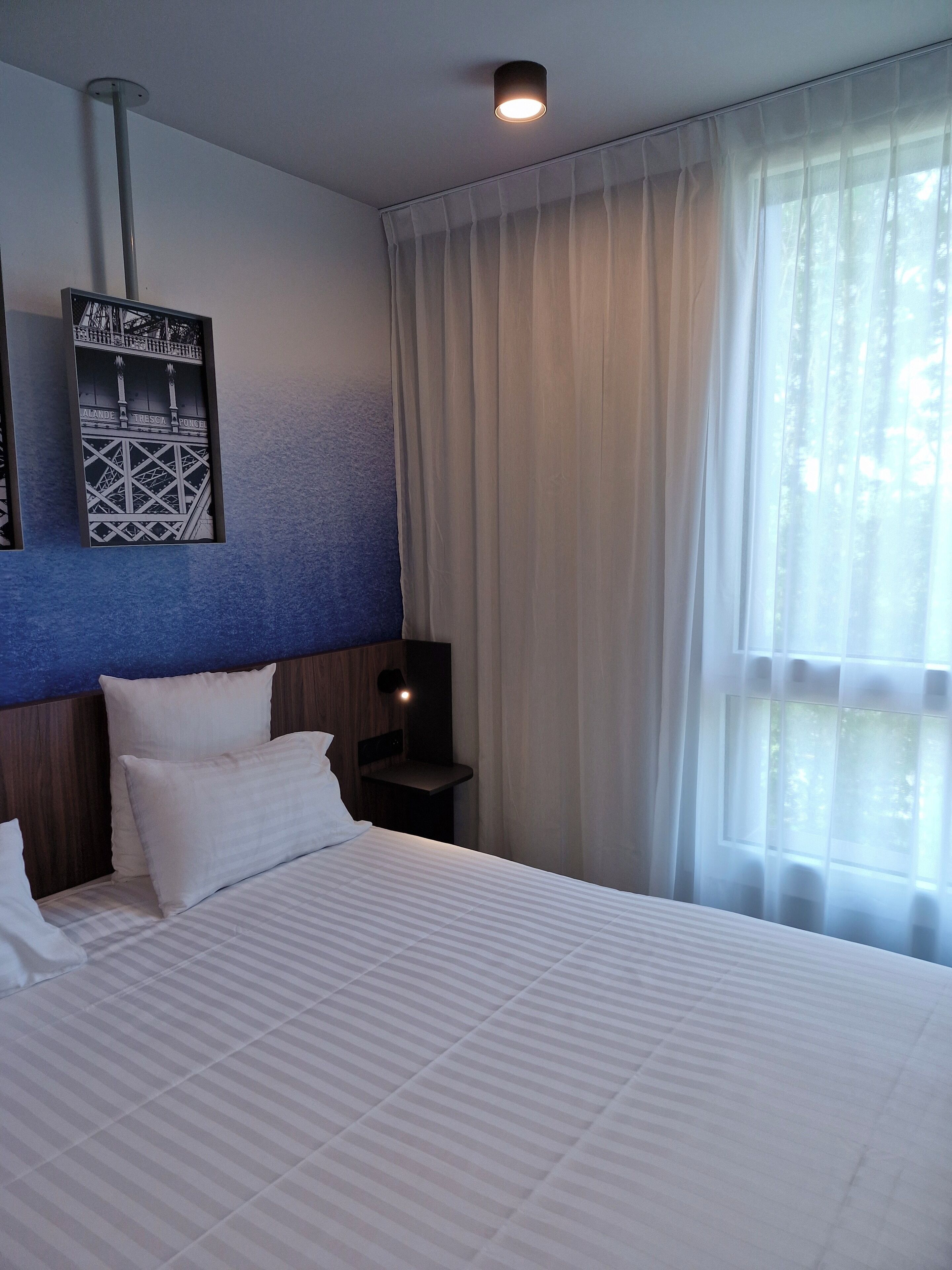 A hotel room with a bed, a headboard with a cityscape design, a nightstand, a wall-mounted light, and a window with sheer curtains.
