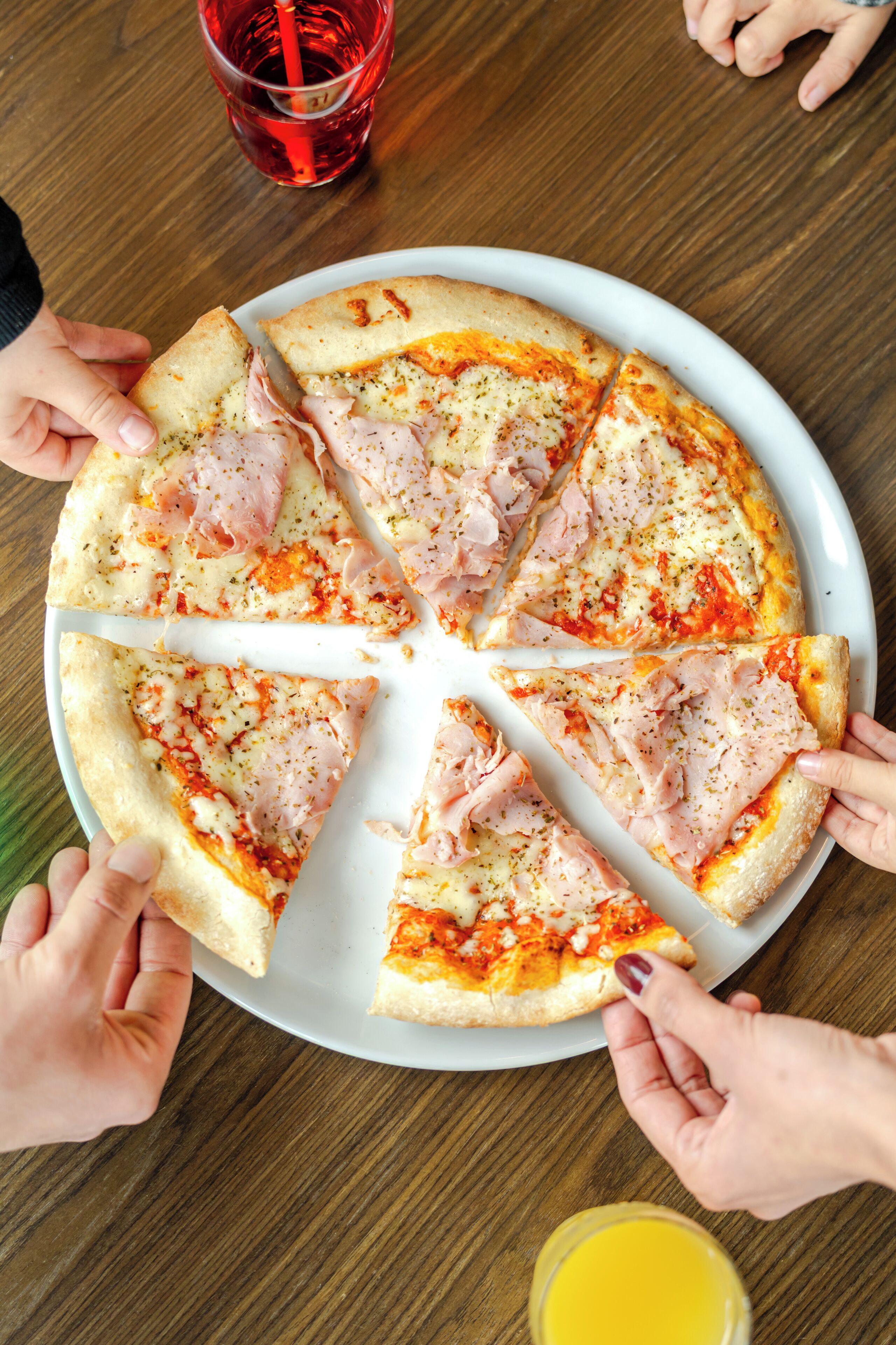 A pizza with ham slices on a white plate, surrounded by people.