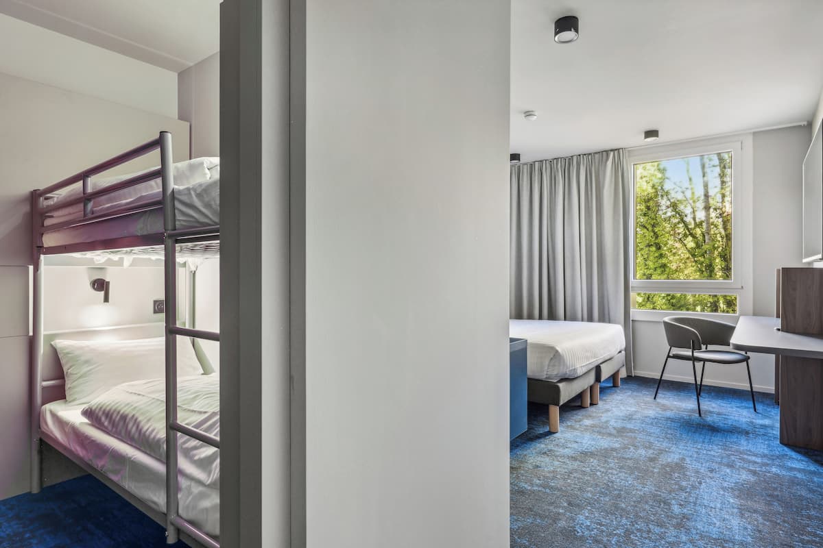 A hotel room with bunk beds, a desk, a chair, and a large window with a view of trees.