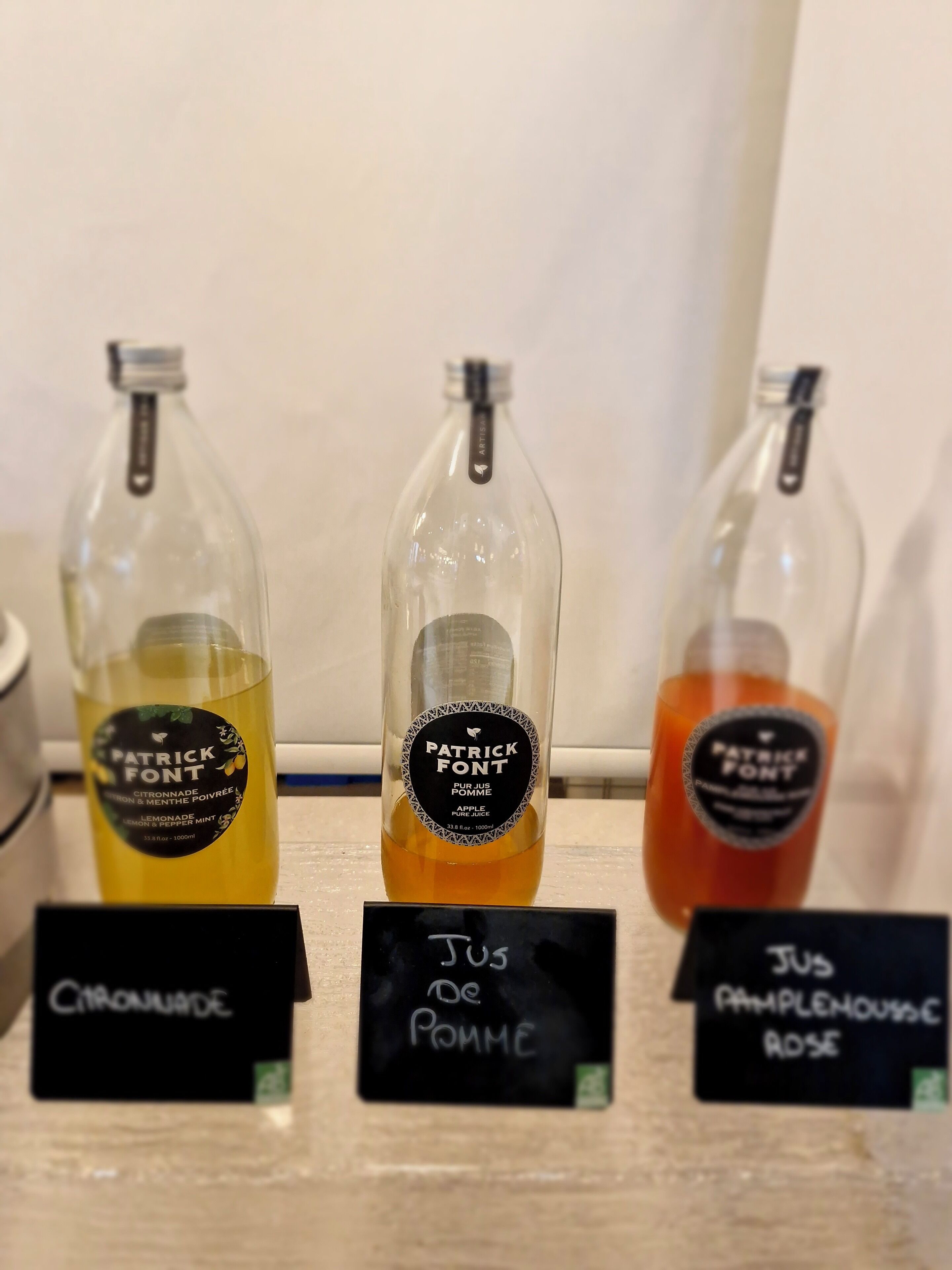 Three bottles of Patrick Font juice with labels indicating flavors: citronnade, pomme, and pomme rose. Each bottle has a black label with white text and a logo.
