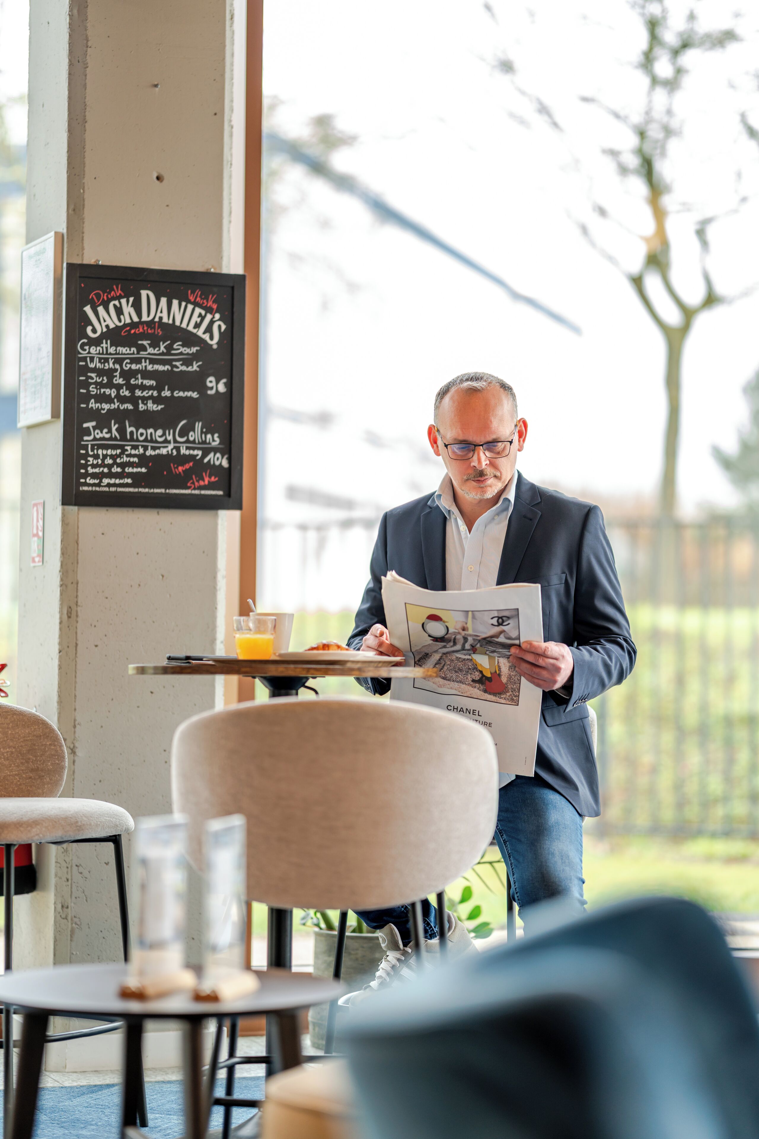 Un hombre leyendo una revista en un café, con una pizarra de menú al fondo.