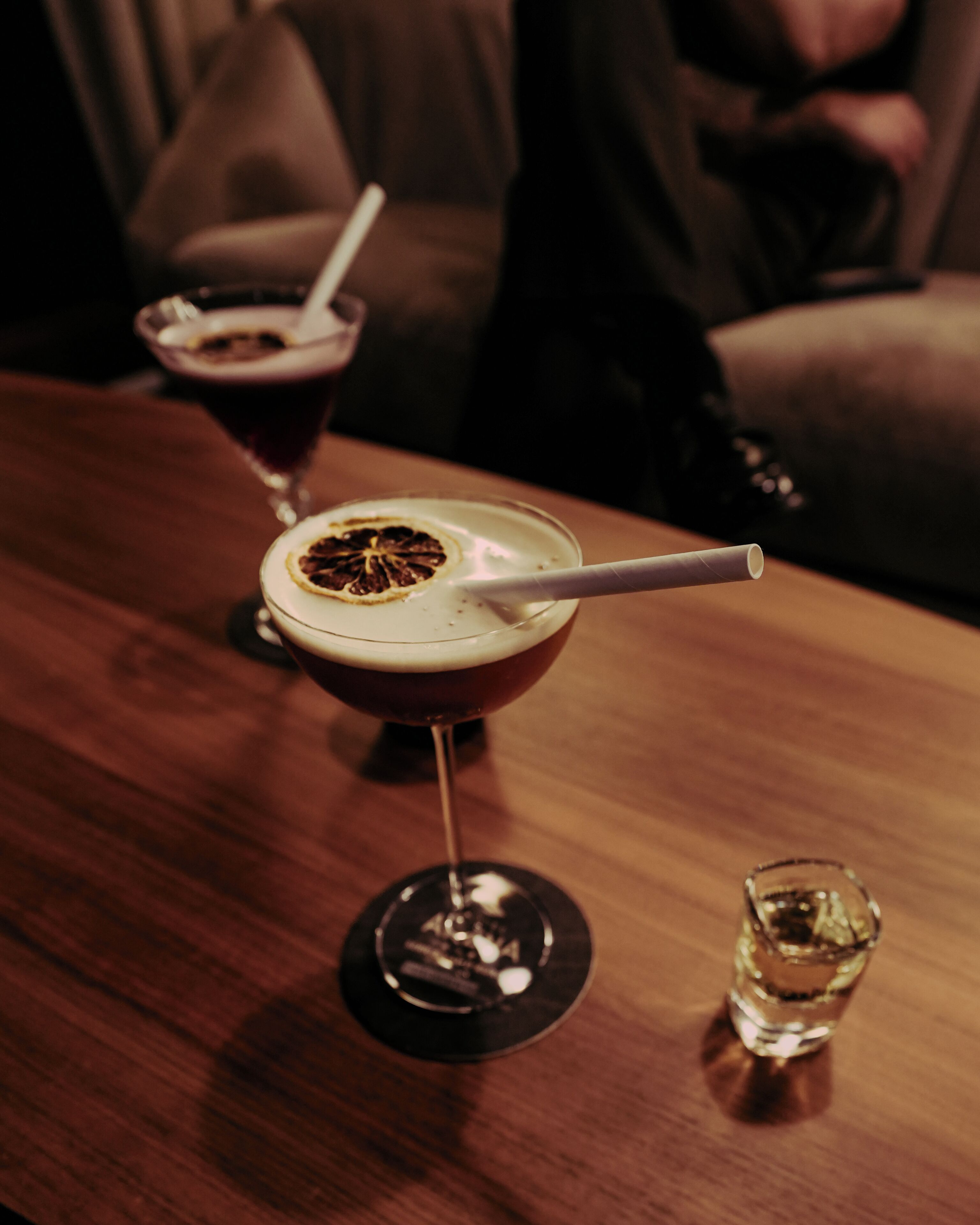A bar setting with a drink in a glass, a shot glass, and a bottle on the table.