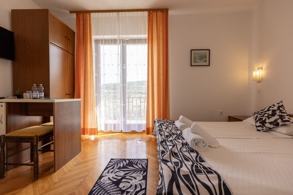 Quarto de hotel com cama, sofá, escrivaninha com cadeira, janela com cortinas e luminária fixada na parede.