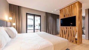 Et moderne hotelværelse med en stor seng, et fjernsyn monteret på en træpanel, en balkon med gardiner, og et badeværelse, der kan ses gennem en åben dør.