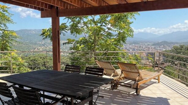 Un espace extérieur abrité, doté d’une table et de chaises, offrant une vue sur le paysage urbain et les montagnes.