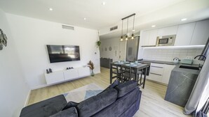 Una sala moderna con un sofá gris oscuro, un televisor de pantalla plana montado en la pared, un comedor con mesa y sillas, y una cocina con microondas y armarios.