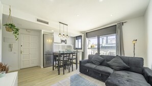Una sala de estar moderna con un sofá gris, una mesa de comedor con sillas y una zona de cocina con refrigerador y microondas.