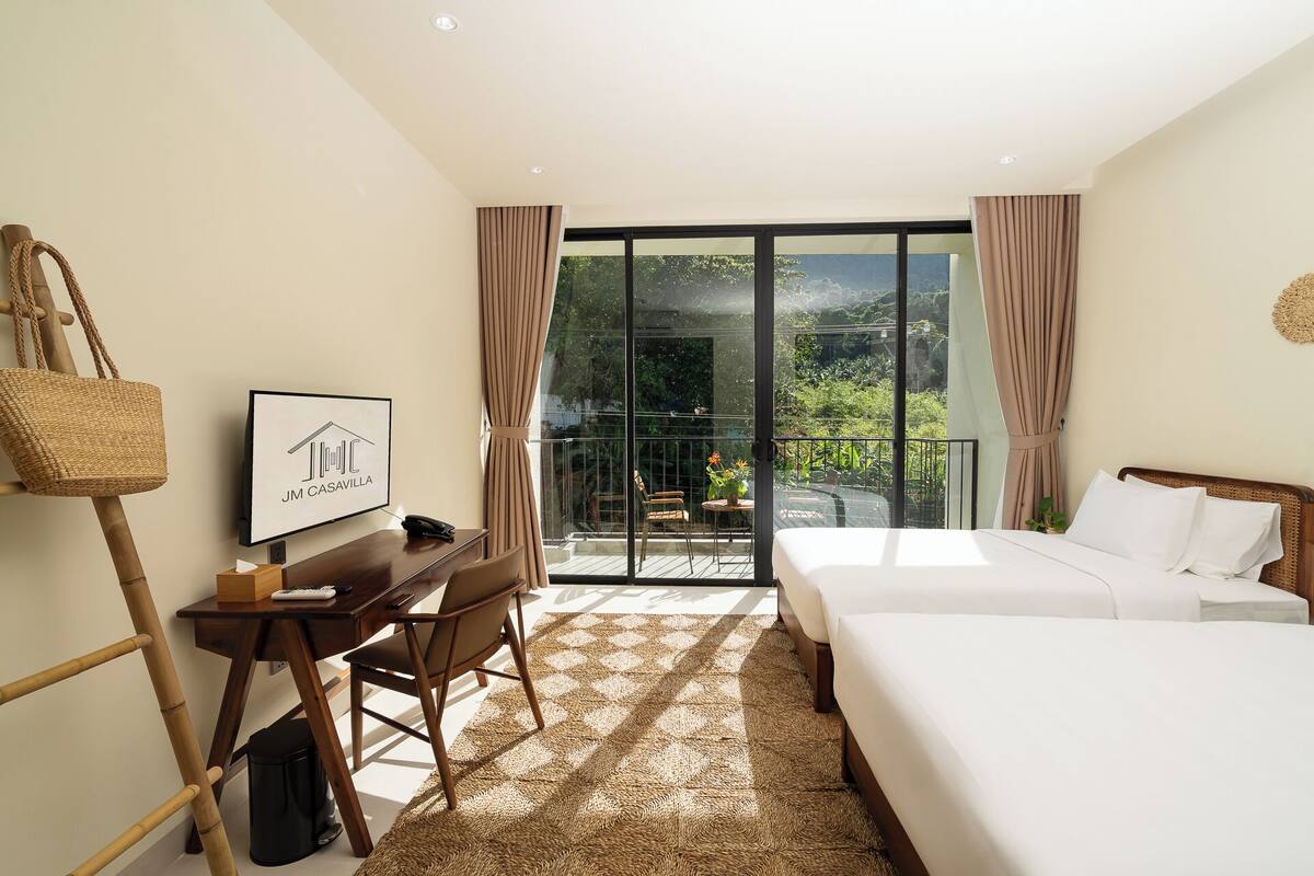 Ein Hotelzimmer mit Bett, Schreibtisch mit Stuhl, Fernseher und einem Balkon mit Blick auf Grünflächen.