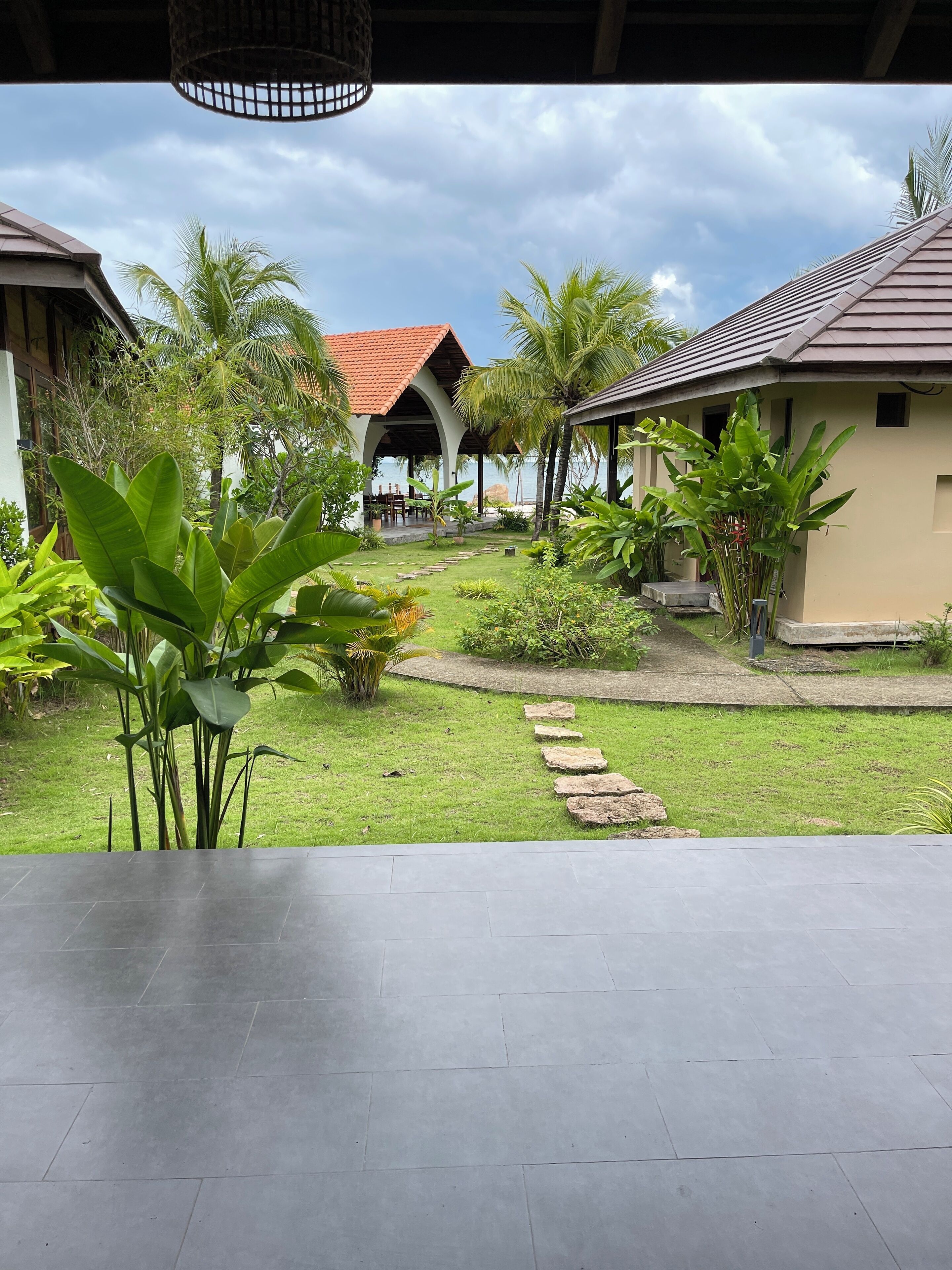 A covered outdoor area with a paved floor, surrounded by lush greenery and palm trees, leading to a building with a red-tiled roof.