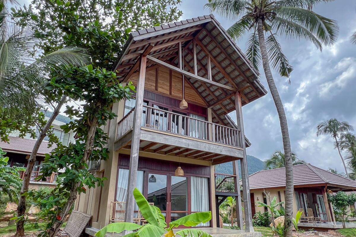 Ein zweistöckiges Holzhaus mit Balkon, umgeben von tropischer Vegetation und Palmen.