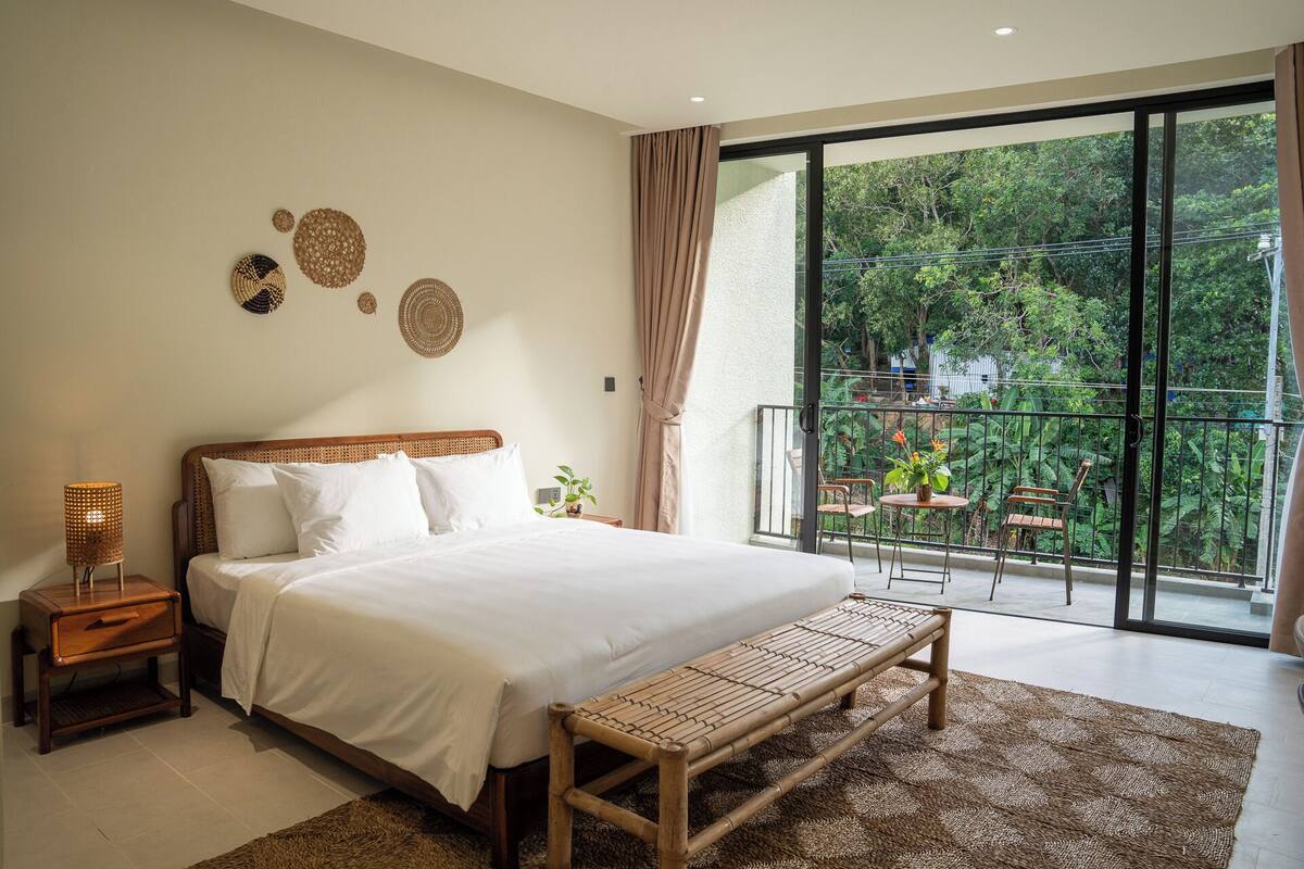 Ein Schlafzimmer mit einem großen Bett, einem Nachttisch, einer Bank und Blick auf einen Balkon mit Gartenmöbeln.