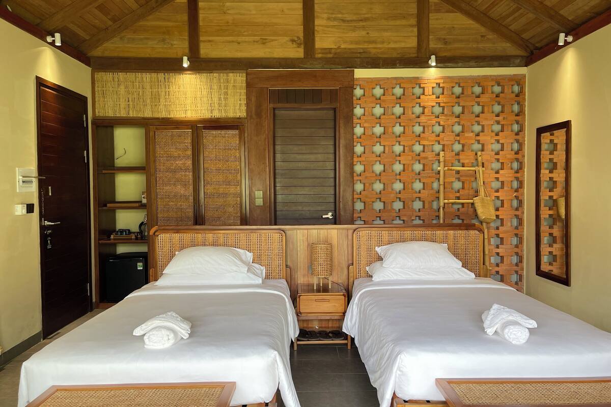 Ein Zimmer mit zwei Betten, Holz-Kopfteilen, einer gewebten Wandverkleidung, einer Tür und einem kleinen Tisch.