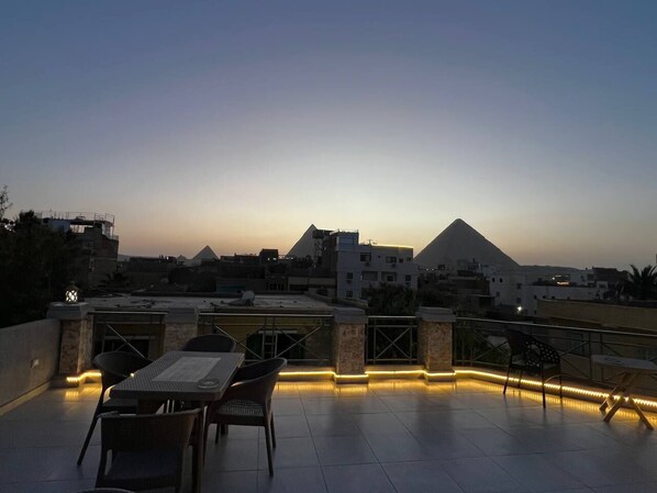 A rooftop terrace with tables and chairs, a pyramid in the background, and a cityscape.