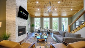 A spacious living room with a stone fireplace, a large TV mounted on the wall, a wooden ceiling, and a staircase to the right.