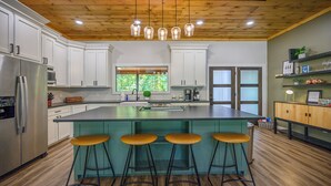 A modern kitchen with white cabinets, a large island with stools, a stainless steel refrigerator, and a wooden ceiling.