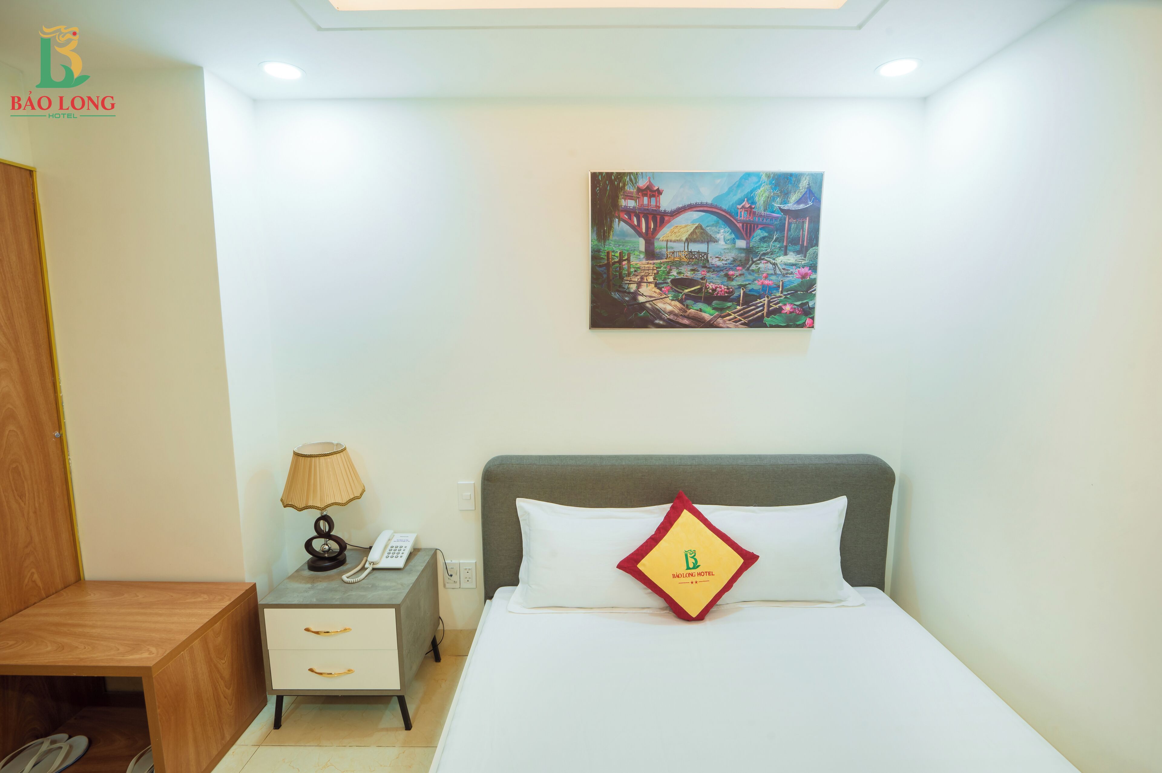 Habitación de hotel con cama, mesita de noche, lámpara, un cuadro en la pared y el logo en la almohada.