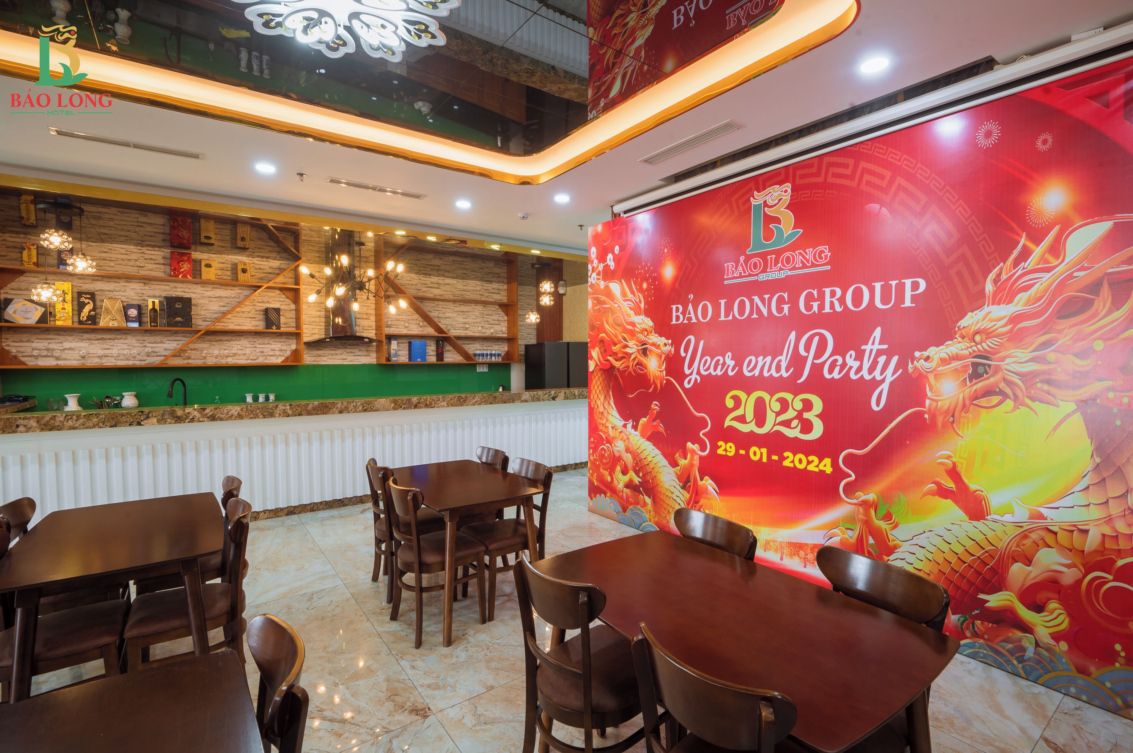Zona ristorante con tavoli e sedie in legno, un bancone con sfondo verde e un grande striscione rosso che pubblicizza una festa di fine anno.