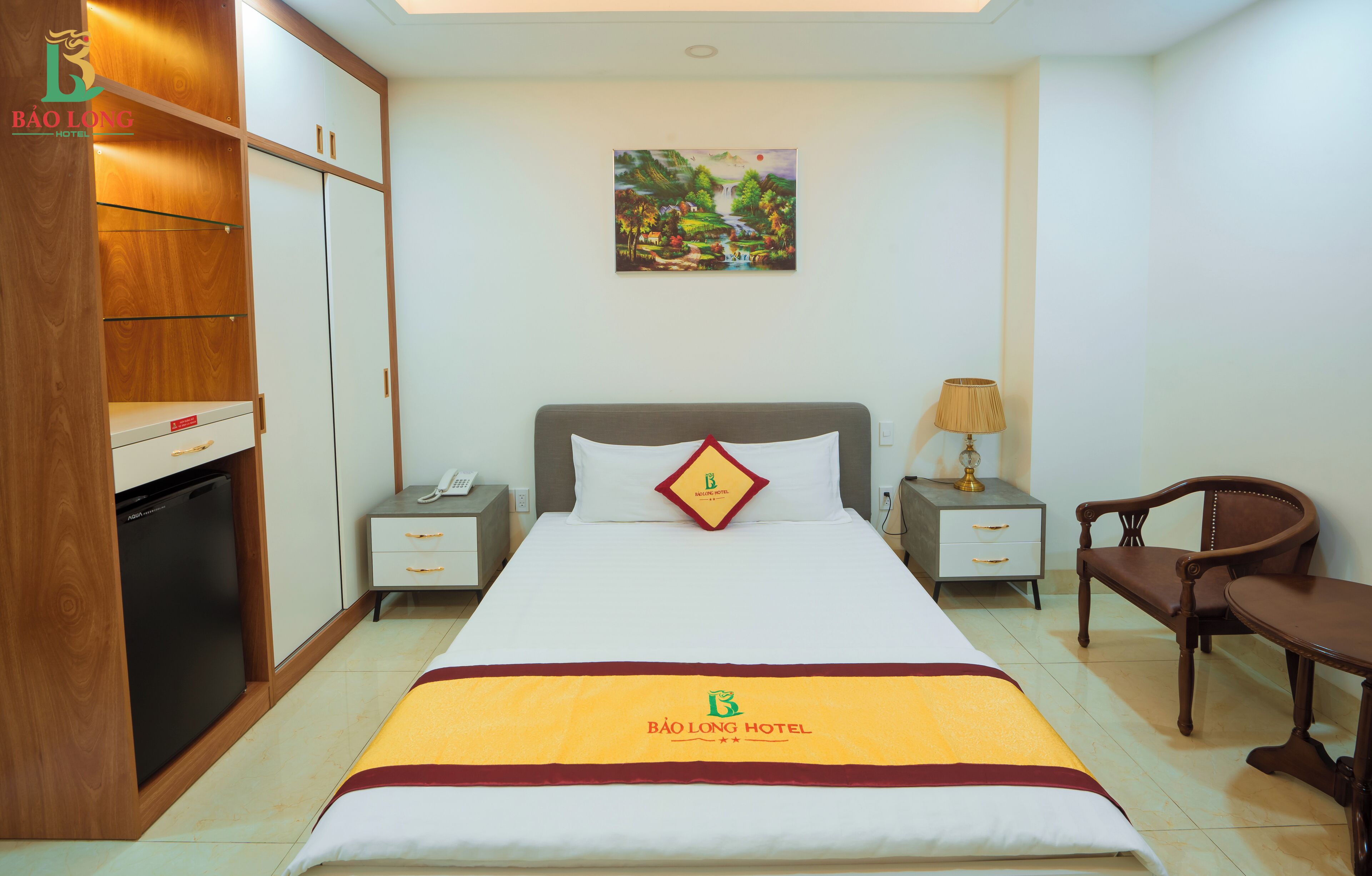 A hotel room with a bed, bedside tables, a desk, a chair, a wardrobe, and a painting on the wall.