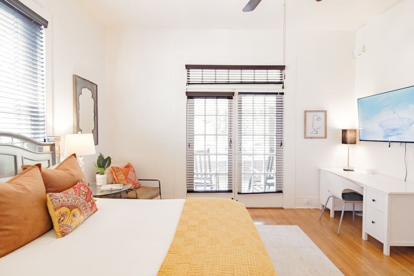 A hotel room with a bed, desk, chair, TV, and large windows with blinds.