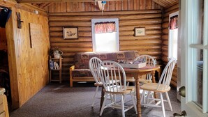A cozy cabin interior with a wooden dining table, chairs, a sofa, and a window with curtains. - Rustic cabin getaway on the shore of Soo Lake in Phillips, WI (Phillips)