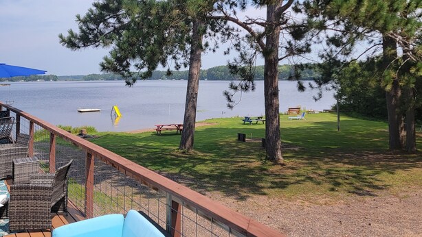 A deck with a blue sofa overlooking a lake, surrounded by trees and picnic tables. - Rustic cabin getaway on the shore of Soo Lake in Phillips, WI (Phillips)