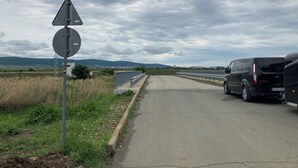A rural road with a van parked on the side, surrounded by fields and distant hills.