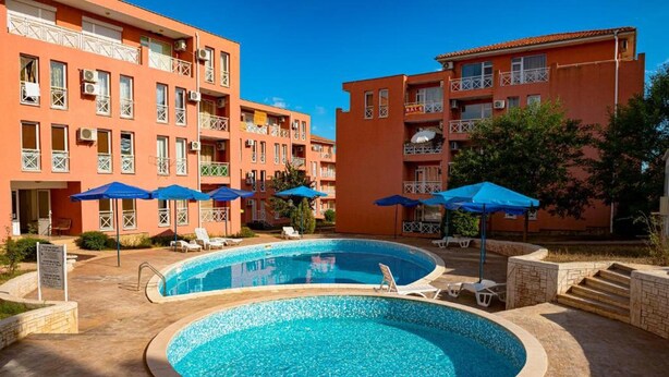 A swimming pool with lounge chairs and umbrellas, surrounded by multi-story buildings with balconies.