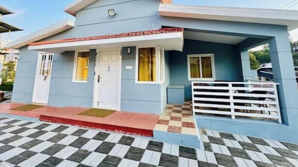 A small, single-story house with a flat roof, a front porch with a white railing, and a checkered tile floor.