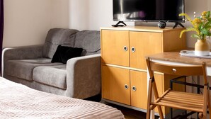 Ett hotellrum med en grå soffa, ett träskåp med en platt-TV, ett litet träbord med en vas med blommor och en trästol.