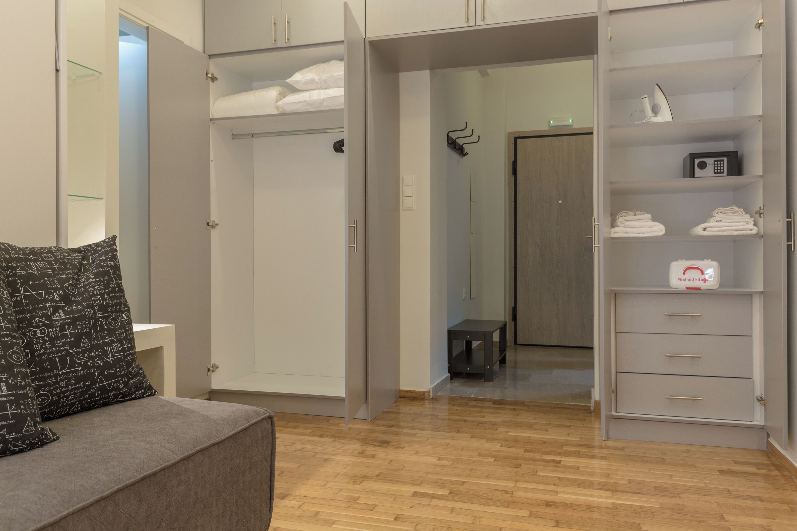 A hotel room with a grey sofa, a wardrobe with multiple compartments, and a small table.