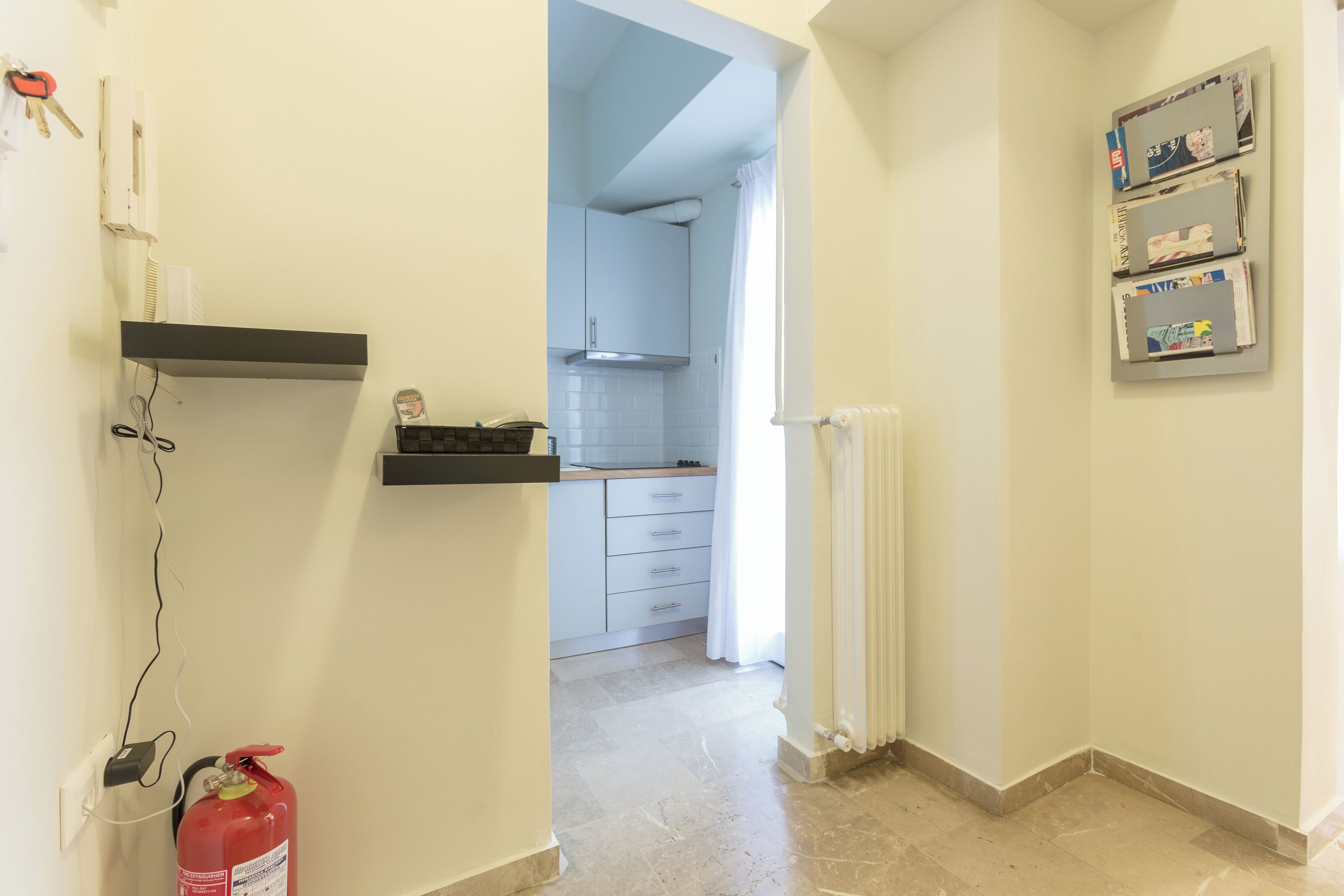 A hallway leading to a kitchen with a fire extinguisher, a radiator, and a shelf with magazines.