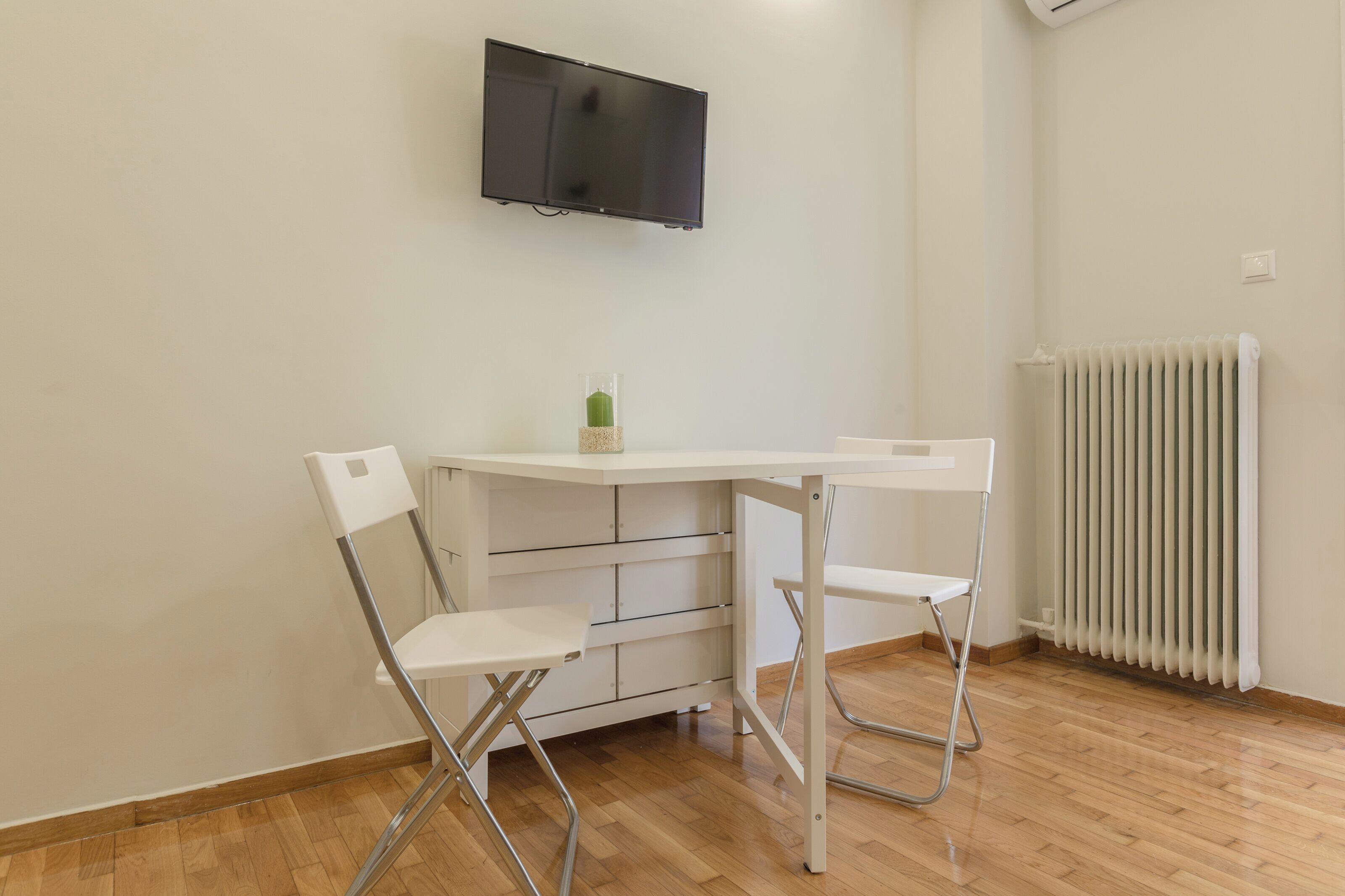 A room with a white table and two white chairs, a mounted TV, and a radiator.