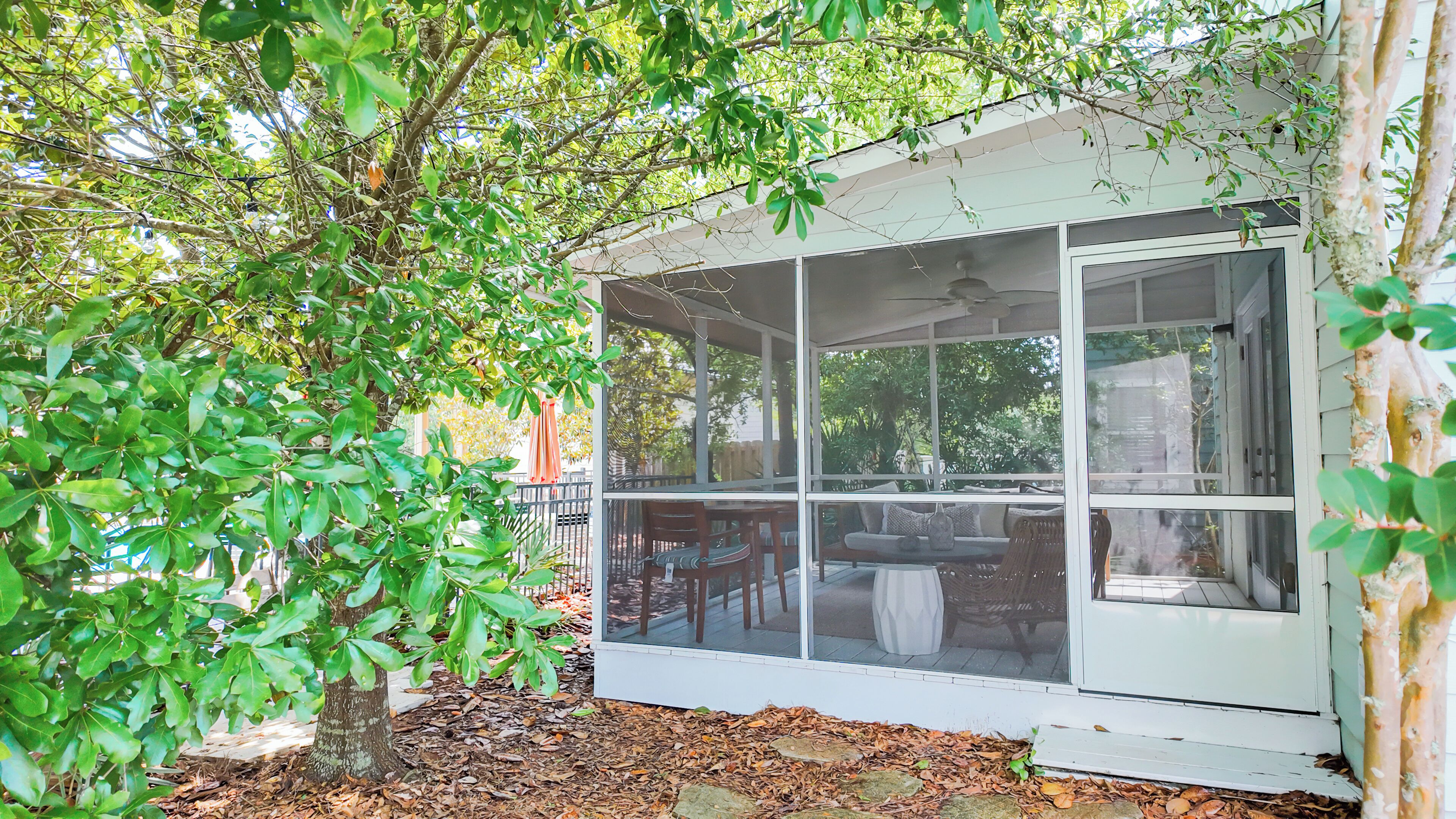 A covered outdoor seating area with a table and chairs, surrounded by trees and a white exterior wall.