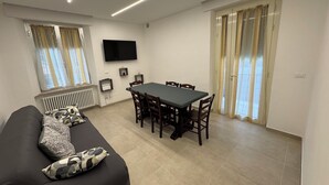 Un salon moderne avec un canapé noir, une table à manger avec des chaises, un téléviseur à écran plat fixé au mur et une porte-fenêtre coulissante donnant sur un espace extérieur.