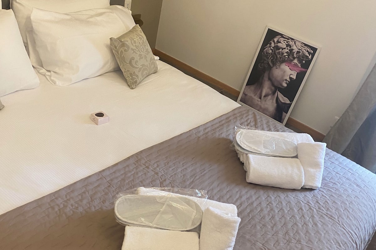 A neatly made bed with white linens, a grey quilted bedspread, and a framed artwork on the wall.