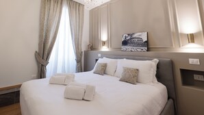 A neatly made bed with white linens and towels, a chandelier, a painting of a building, and a wall-mounted light fixture. - De Vince Luxury Suites - Roma Termini (Rome)