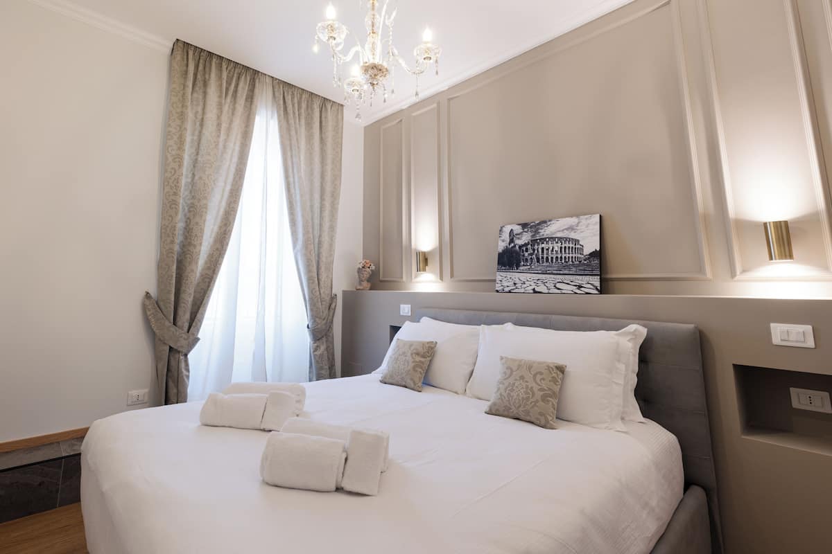 A neatly made bed with white linens and towels, a chandelier, a painting of a building, and a wall-mounted light fixture.