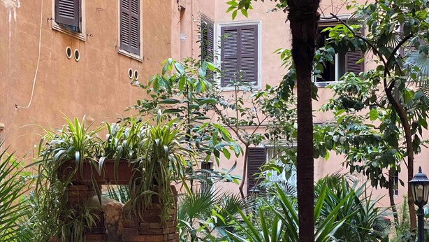 A courtyard with a brick structure, lush greenery, and a building with shutters in the background. - De Vince Luxury Suites - Roma Termini (Rome)